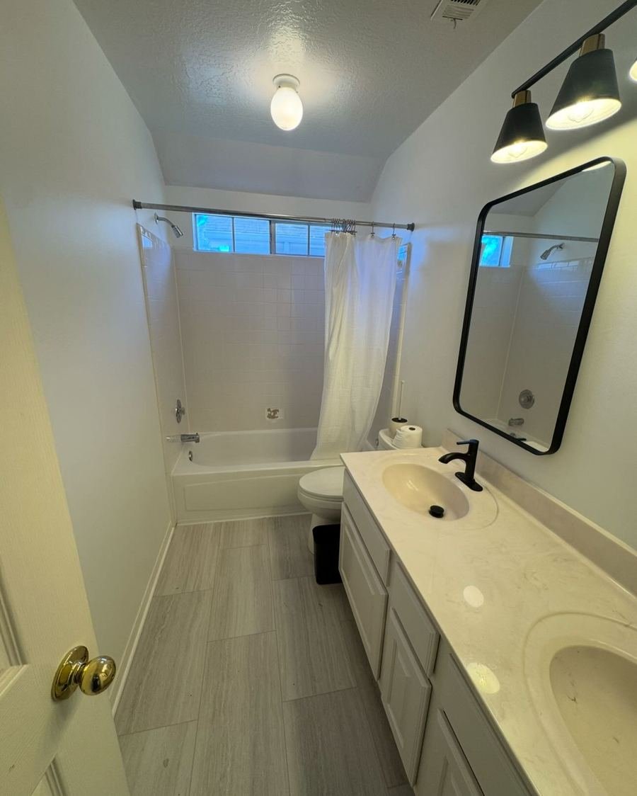 Hall bathroom with gray wood-look floor tile, a white vanity, a black framed mirror and a black three-light vanity fixture