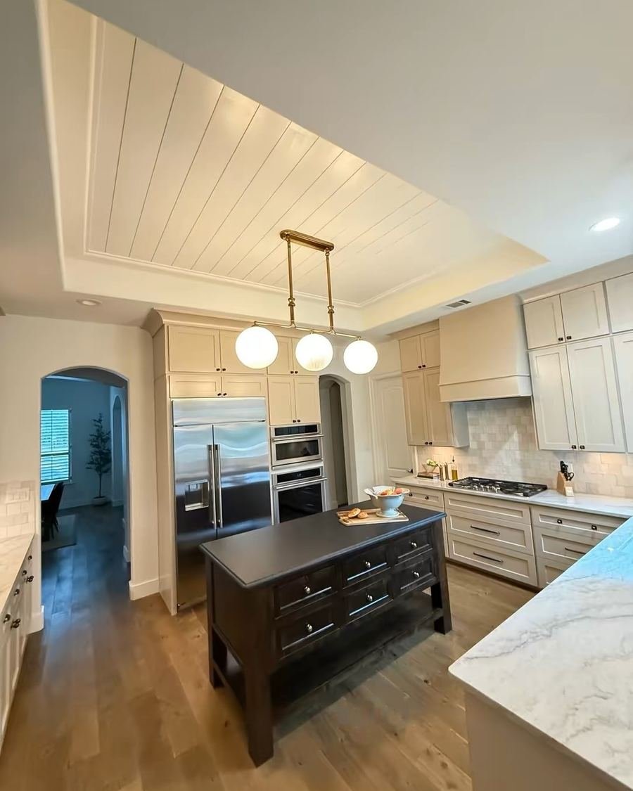 After: the same kitchen with a white planked tray ceiling, a three-globe brass pendant, a dark island with drawers and white veined countertops