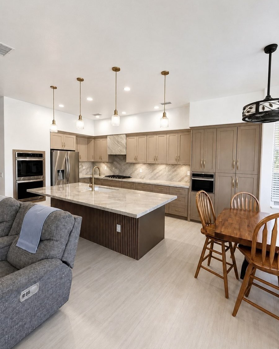 After: the same kitchen with taupe shaker cabinets, a fluted dark island with a light veined top, brass pendants with ribbed glass, a tiled range hood and light wood-look plank floors