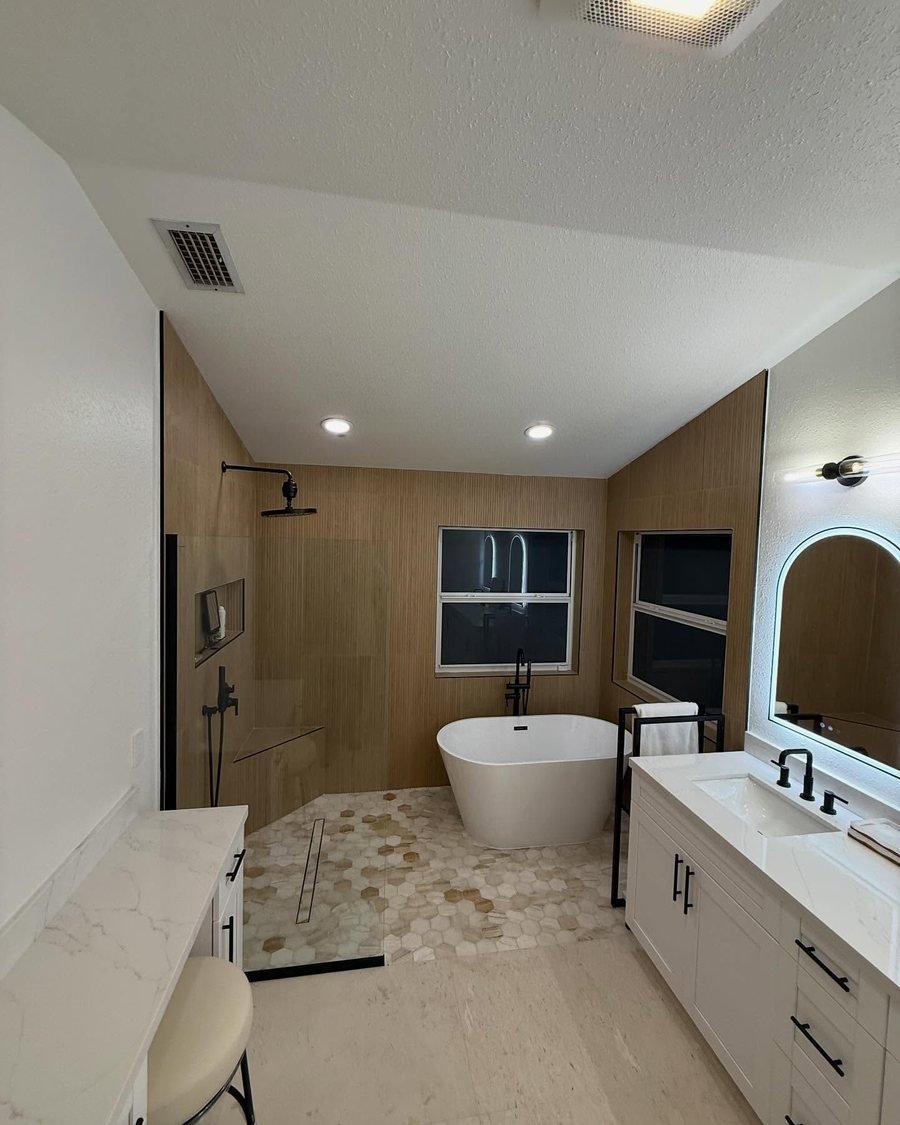 After: the same bathroom with a freestanding tub, a walk-in shower with a glass panel and linear drain, ribbed wood-look wall tile, a hexagon mosaic floor and white vanities with matte black fixtures