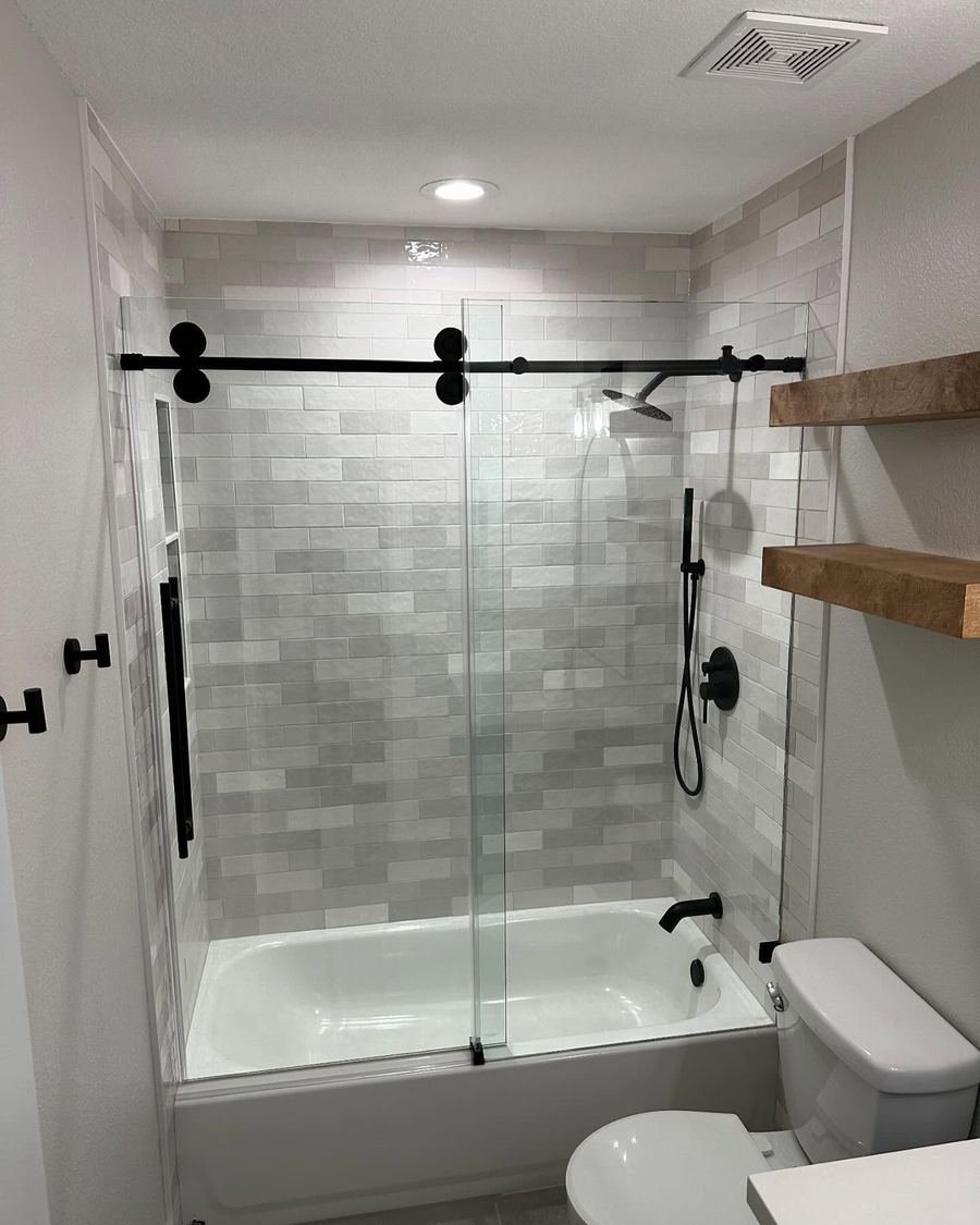 After: the same bathroom with a new alcove tub, gray subway-style tile to the ceiling, a sliding glass door, matte black fixtures and floating wood shelves