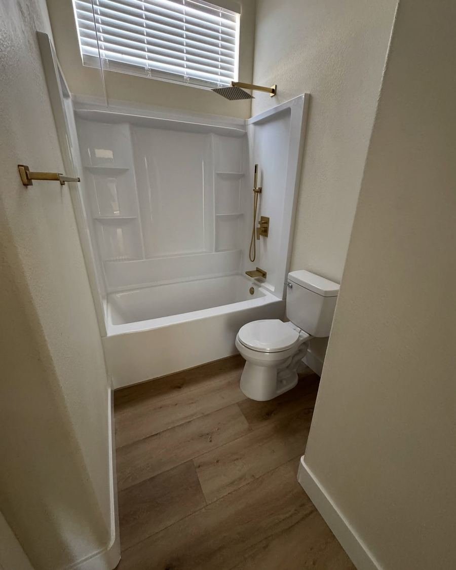 Bathroom with a white tub surround with built-in shelves, gold shower fixtures and wood-look flooring