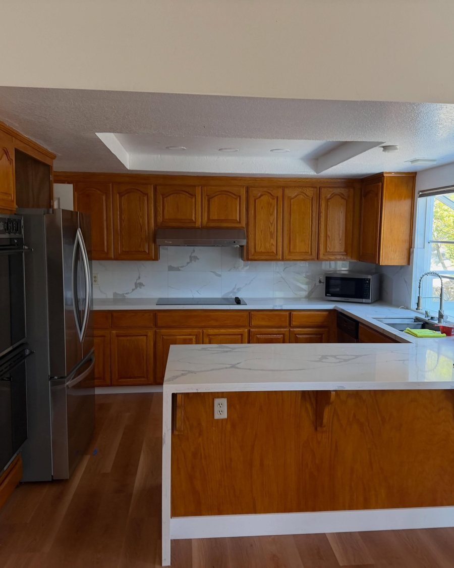 Before: kitchen with honey oak cathedral-style cabinets, white marble-look countertops and a waterfall peninsula