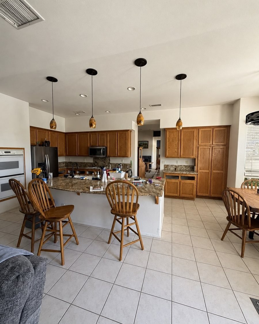 Before: open kitchen with honey oak cabinets, a speckled granite island, amber glass pendant lights and white floor tile