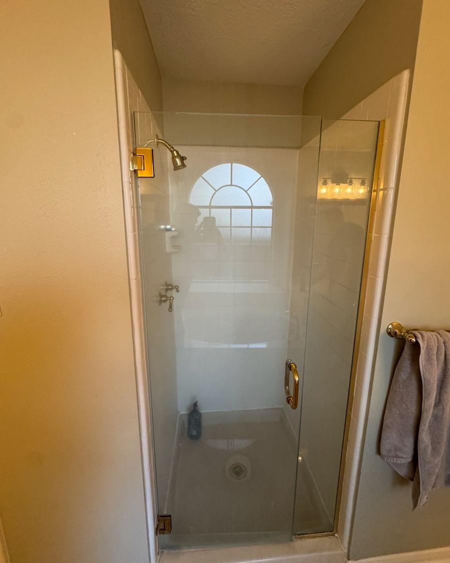 Before: a narrow shower with plain white wall panels and a glass door with brass hardware