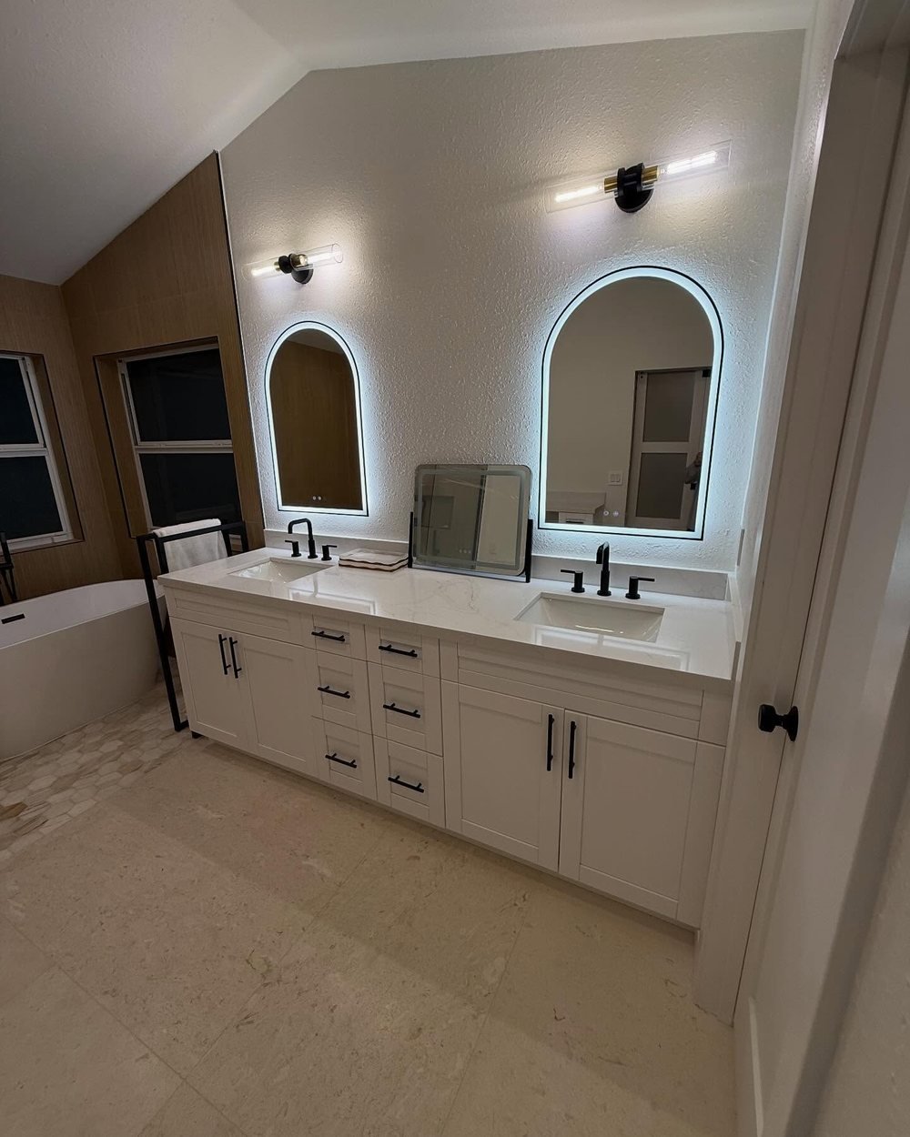 White double vanity with a veined top, matte black faucets, arched backlit mirrors and glass vanity lights